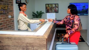 A reception desk with a wooden counter, a digital screen displaying information, and a brick wall with a clock and framed pictures. - Five Clover Hotel Monestery Road (Lekki)