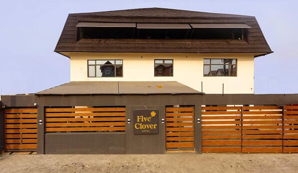 A hotel with a brown roof, a sign reading "Five Clover Hotel," and a wooden fence with horizontal slats. - Five Clover Hotel Monestery Road (Lekki)