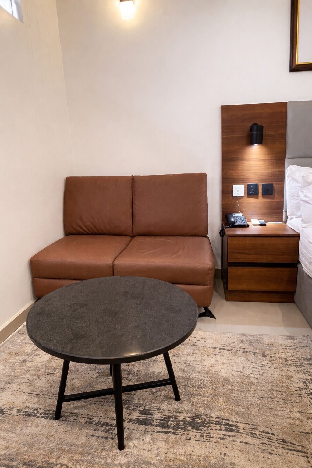 A modern hotel room with a brown sofa, a round table, a wooden nightstand, and a wall-mounted lamp.