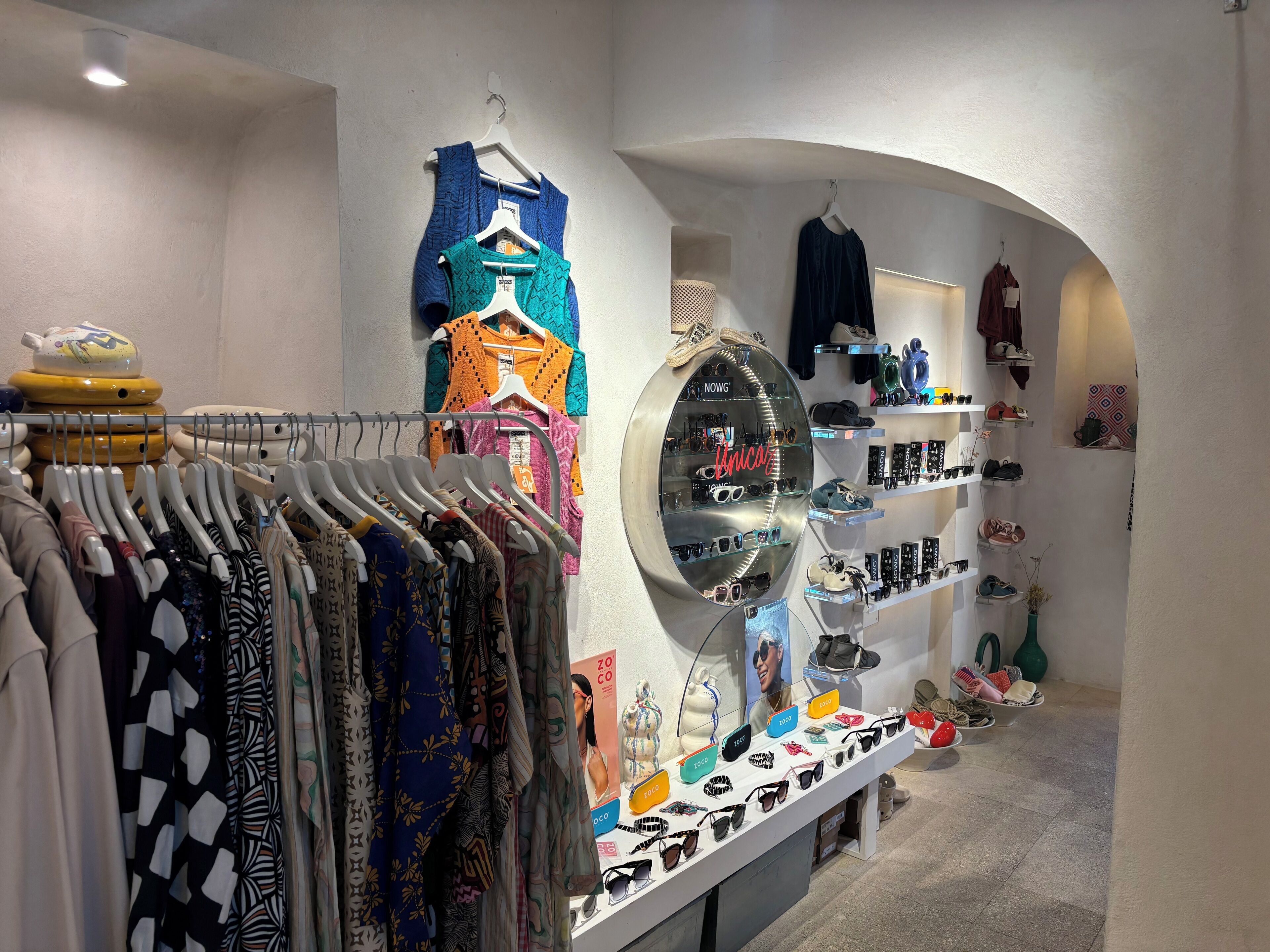 A clothing store with racks of clothes, a display of sunglasses, and shelves with shoes.