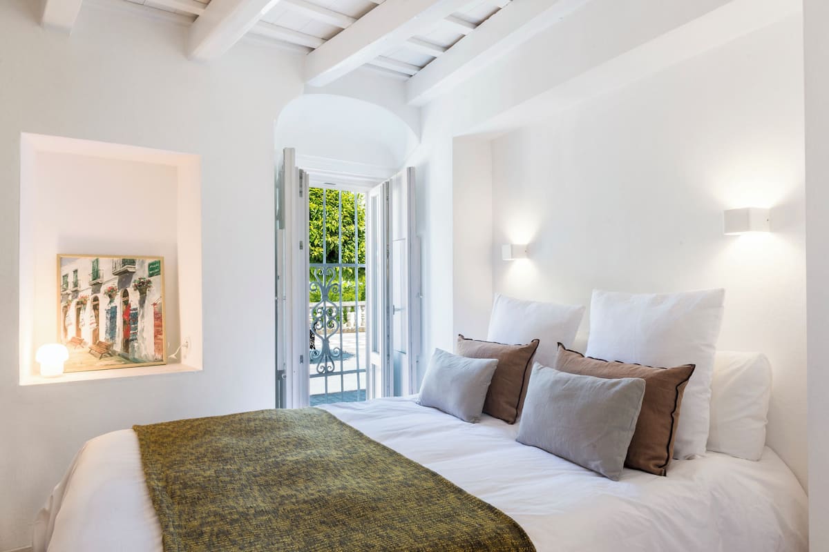 Une chambre à coucher avec un lit, des oreillers, un tableau et une vue sur l’extérieur, visible à travers une porte ouverte.