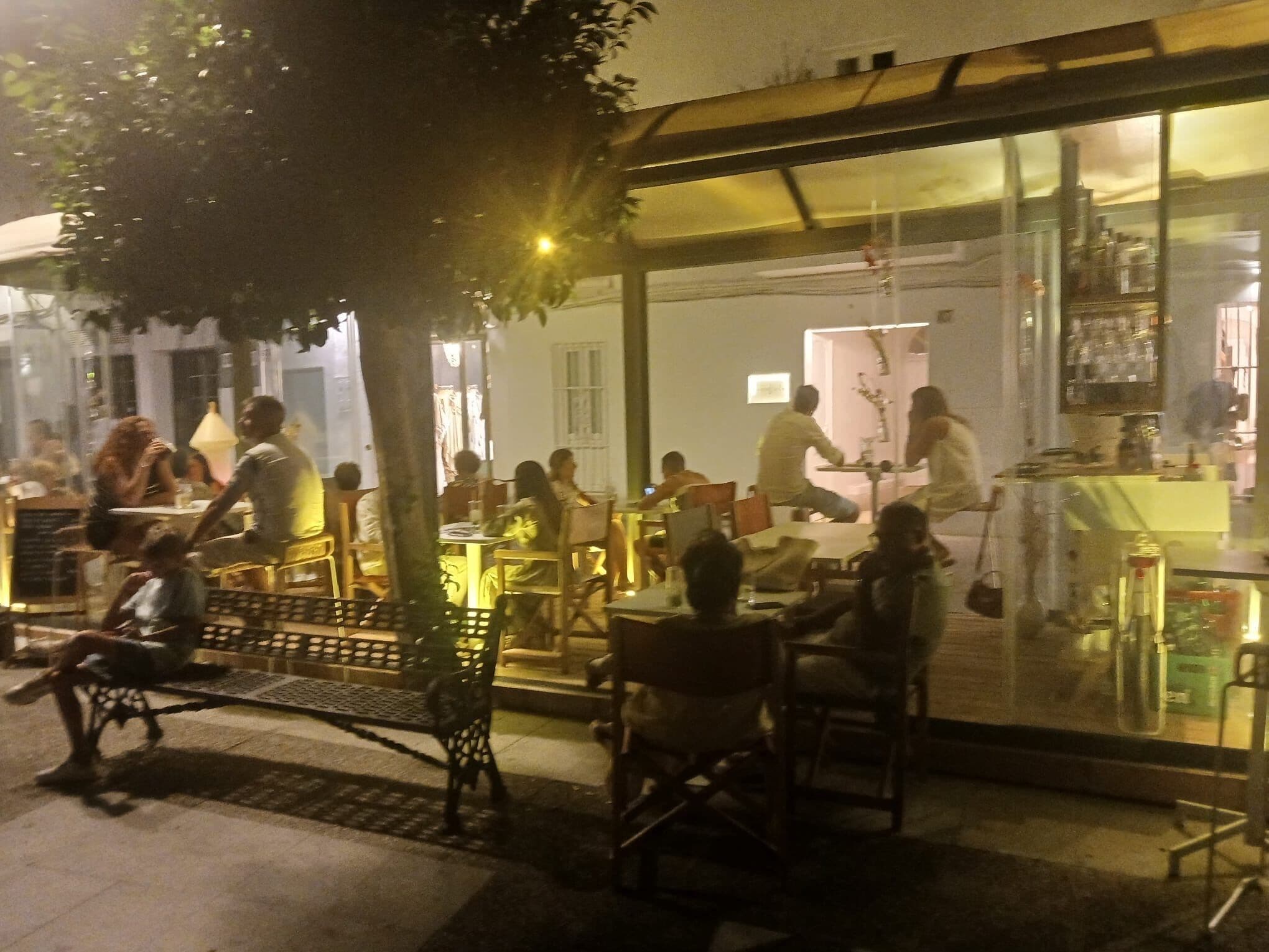 An outdoor dining area with tables and chairs, a tree, and a building with a glass facade in the background.