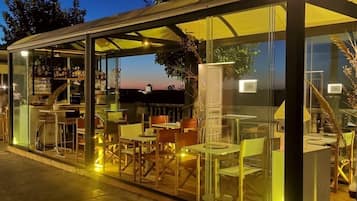 A glass-enclosed outdoor seating area with tables and chairs, a bar with bottles, and a view of the sunset.