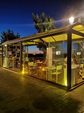 Een overdekte buitenruimte met zitplaatsen, tafels en stoelen, een bar met flessen en uitzicht op de ondergaande zon.