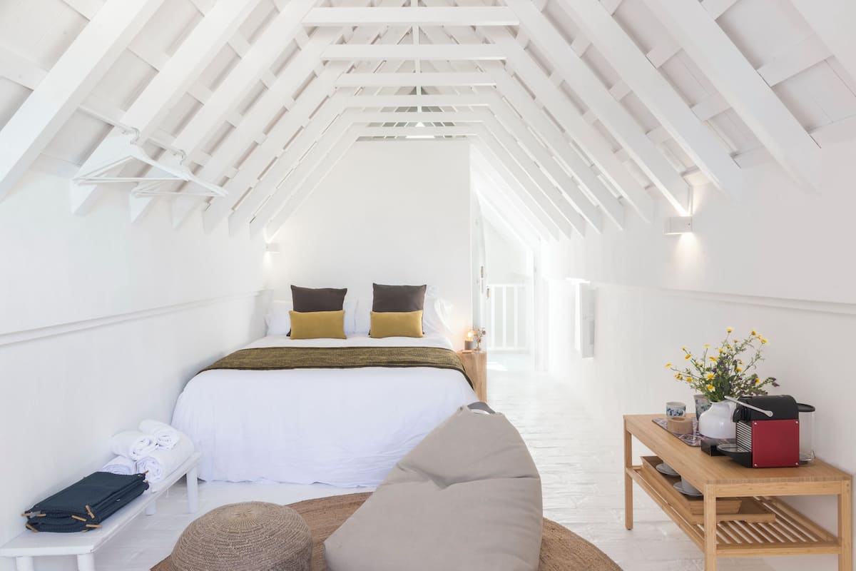Une chambre avec un plafond en pente, un lit avec une literie blanche et deux oreillers, une table de chevet avec une lampe, un banc en bois et une petite table sur laquelle se trouvent une machine à café et des fleurs.