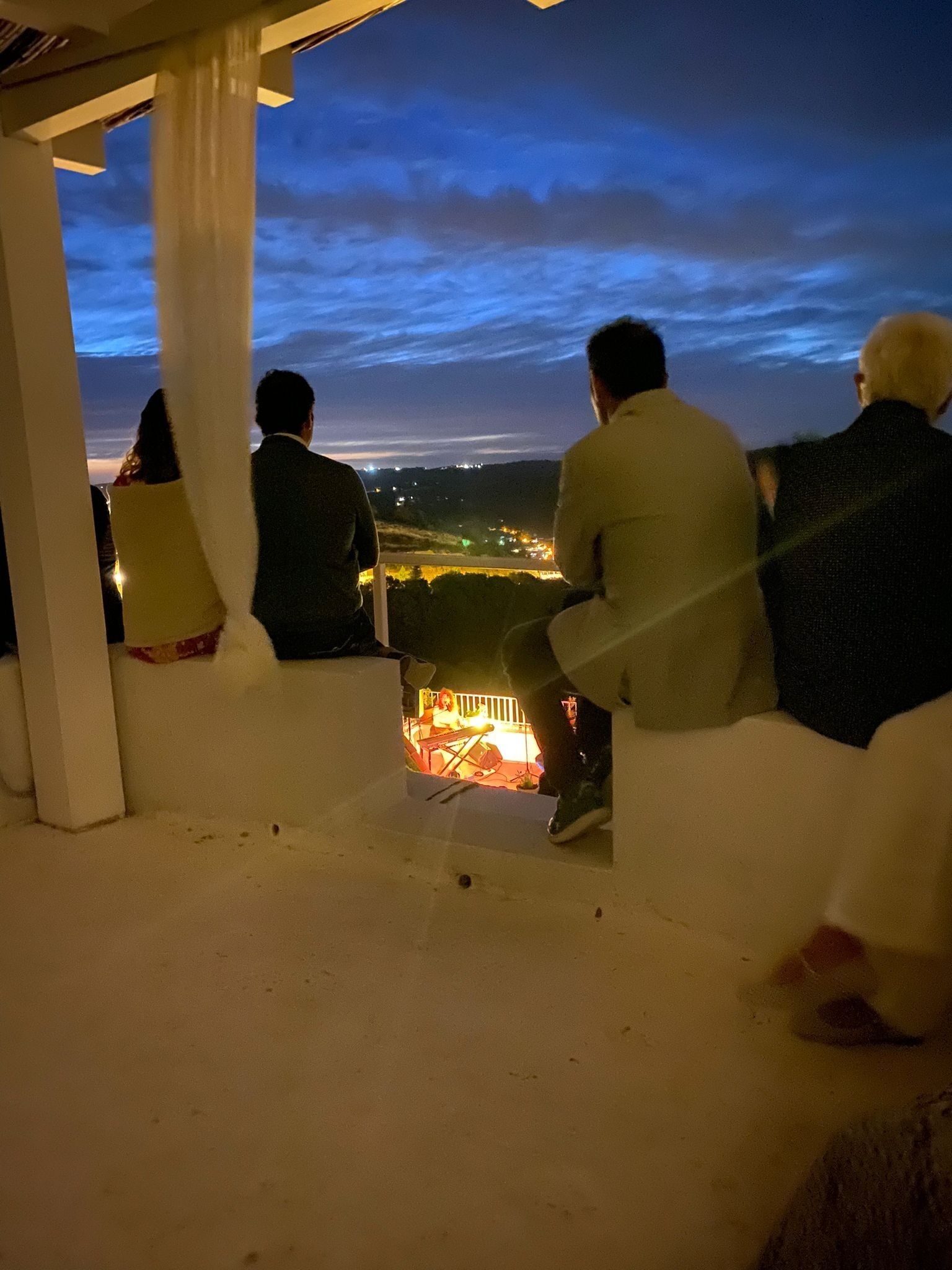 Un grupo de personas sentadas en un banco blanco con vistas a un paisaje urbano al anochecer.