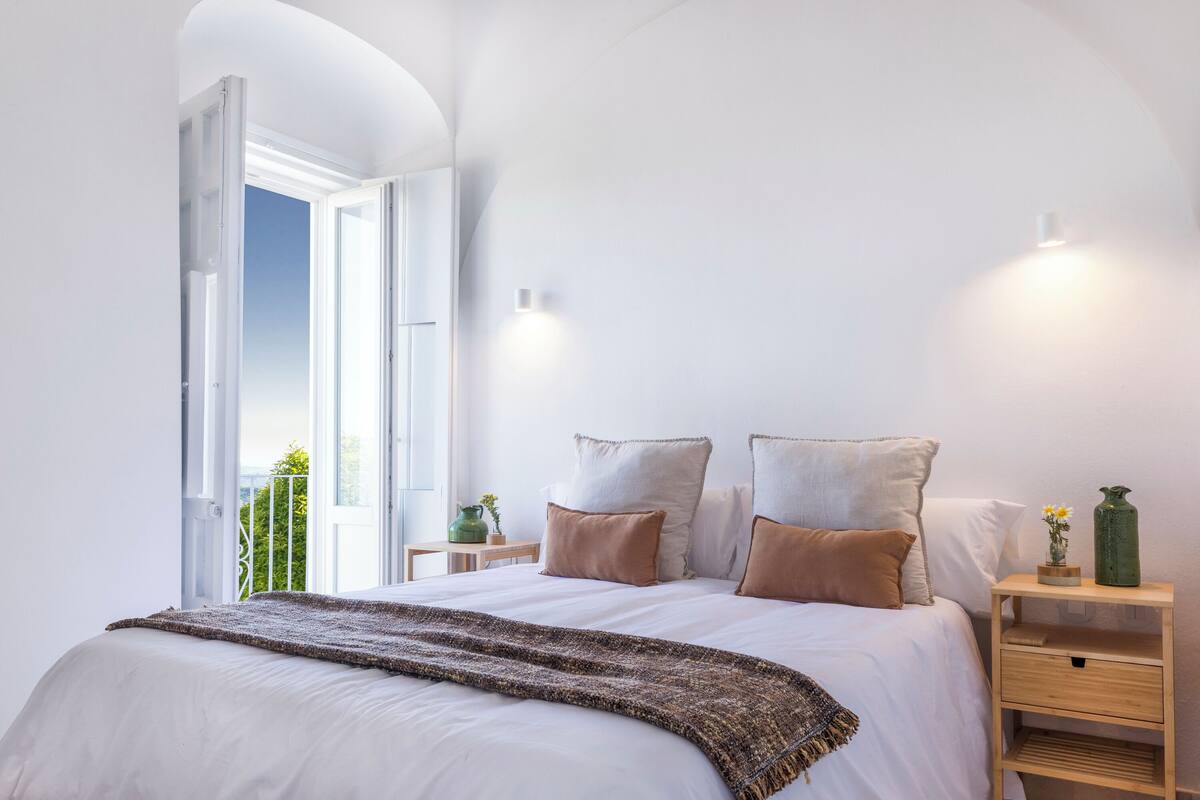 Une chambre à coucher avec un grand lit, une table de chevet, une commode et une fenêtre donnant sur un paysage verdoyant.