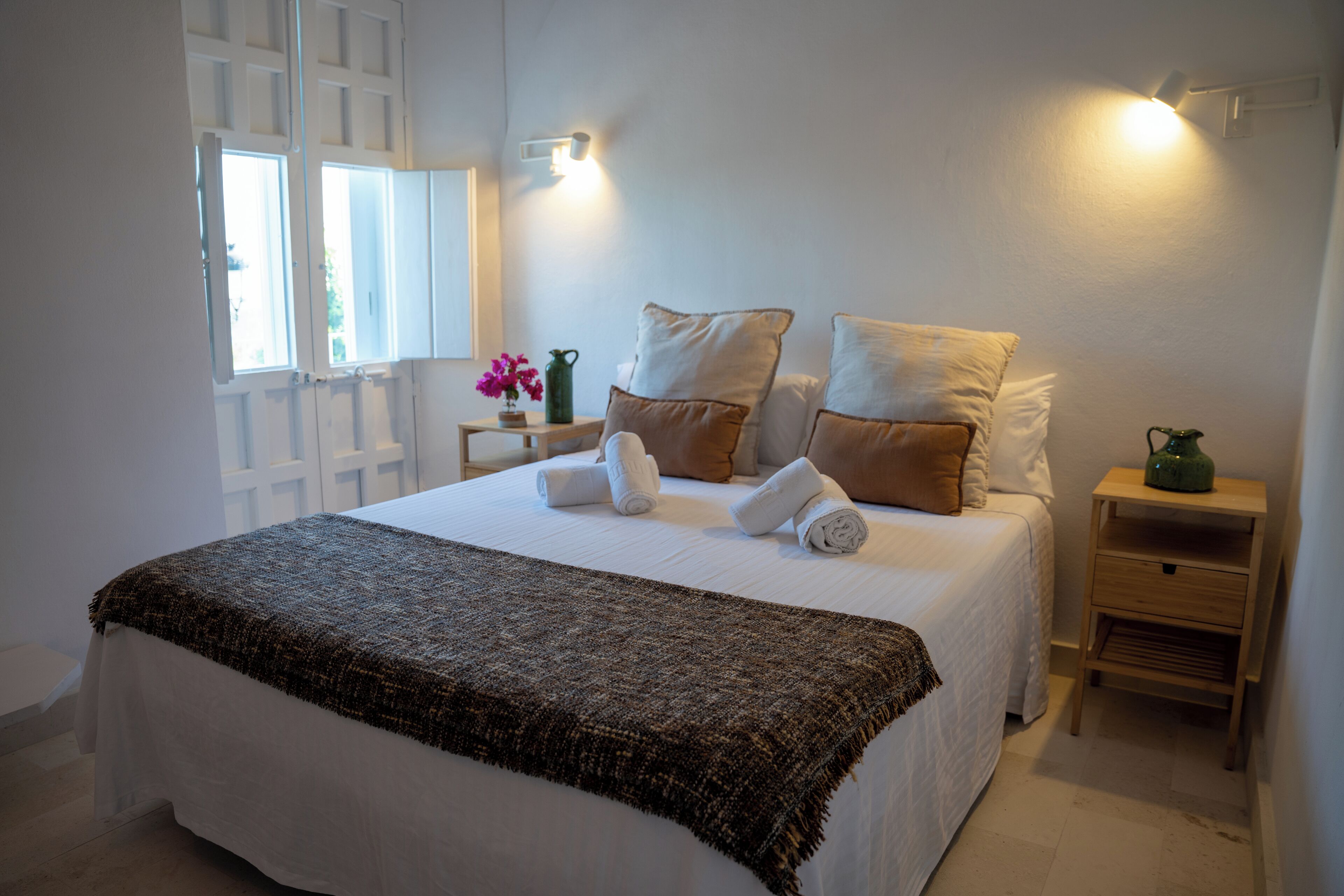 A neatly made bed with a white comforter and a brown throw blanket, a small bedside table with a green vase, and a window with white shutters.
