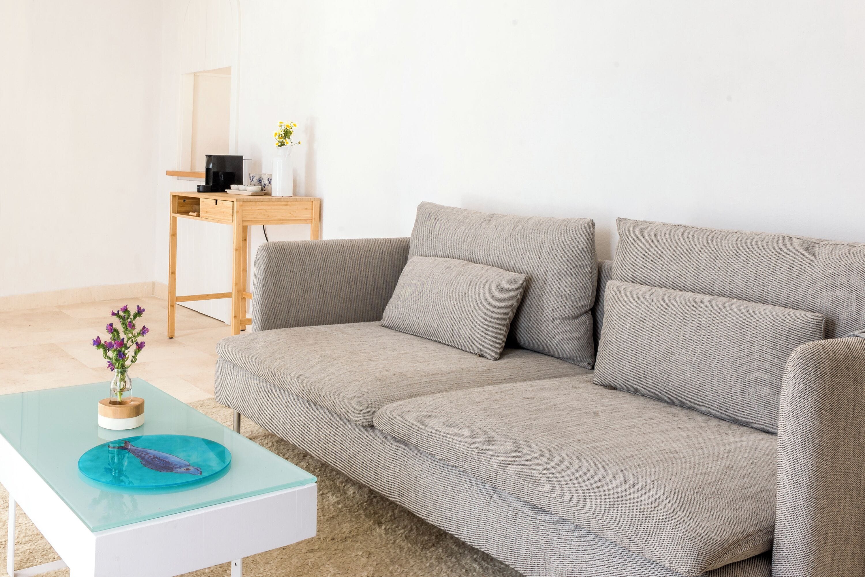 A modern living room with a grey sofa, a glass coffee table, and a wooden side table with a coffee machine and a vase of flowers.