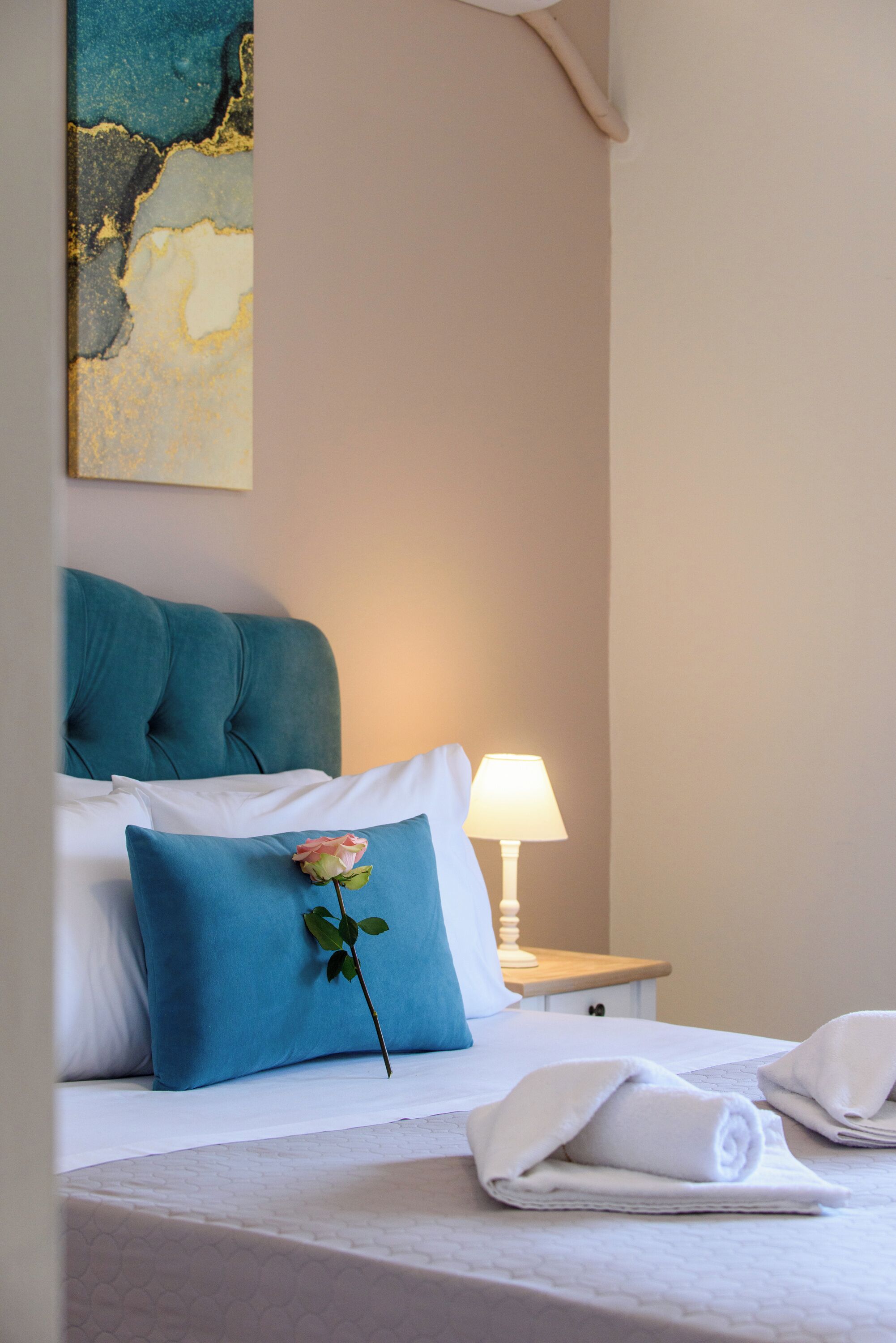 Un lit bien fait, avec une tête de lit de couleur turquoise, des oreillers blancs et une seule rose en guise de décoration.