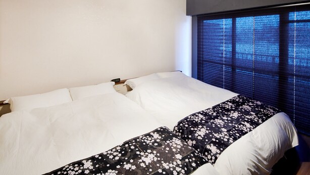 Un dormitorio con cama, ropa de cama blanca, una colcha de flores en blanco y negro, una ventana con persianas y una lámpara colgante.