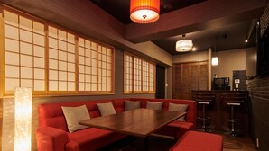 Una habitación tradicional japonesa con suelo de tatami, una mesa de madera baja, cojines rojos, puertas correderas shoji y una zona de cocina con microondas y taburetes de bar.