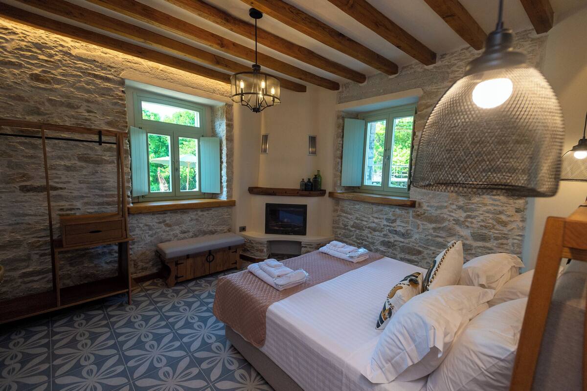 Une chambre confortable avec un mur en pierre, des poutres en bois, un grand lit, un banc et une cheminée.