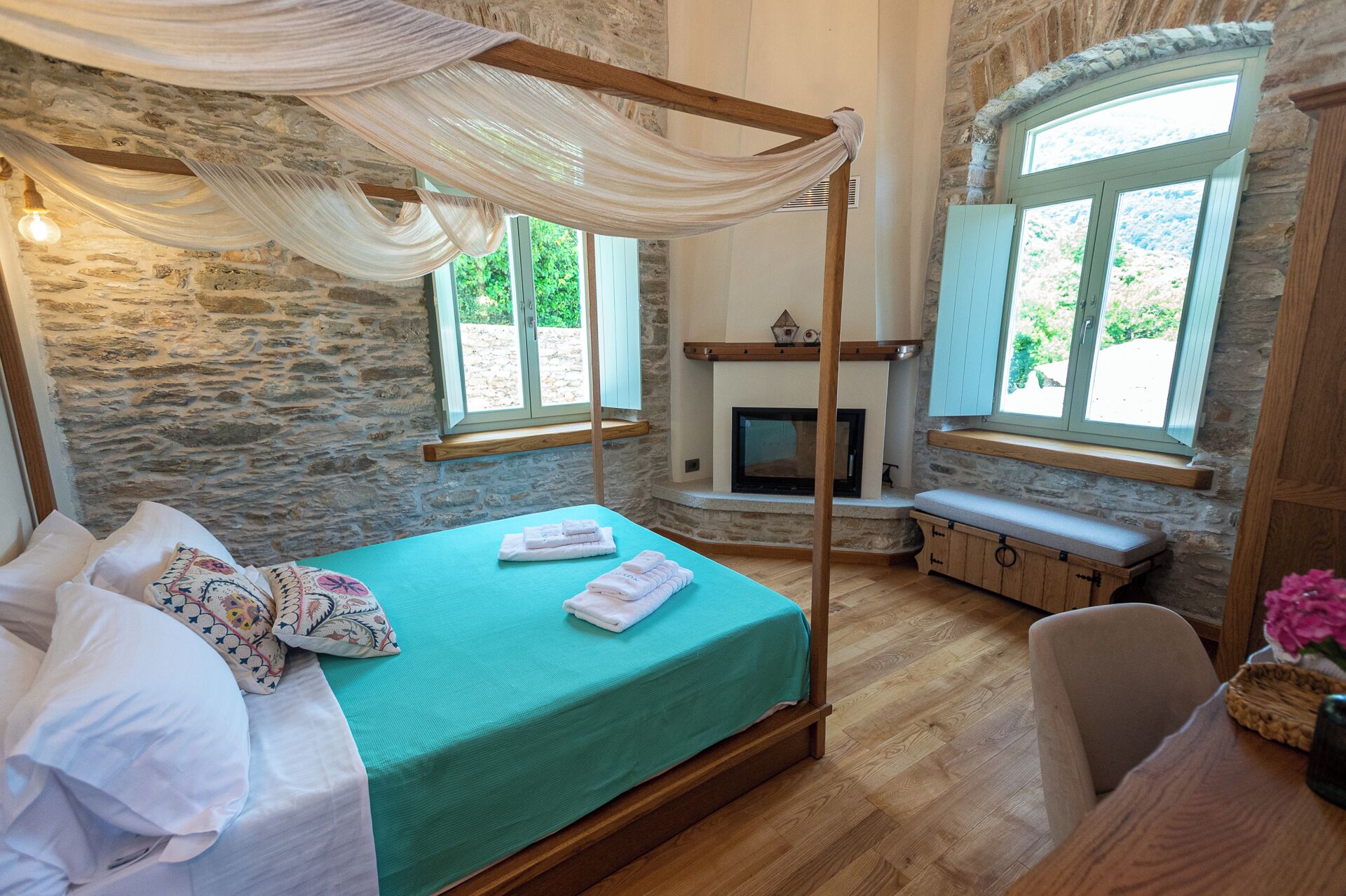 A room with a four-poster bed, a stone wall, a fireplace, and large windows.