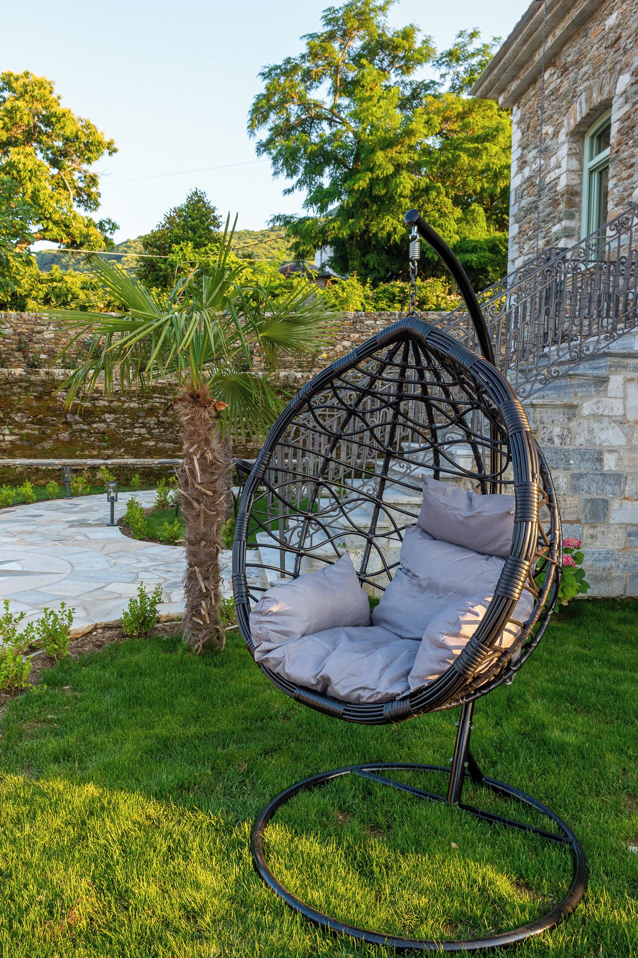 Un fauteuil suspendu en osier, agrémenté de coussins, se trouve dans un jardin, avec un bâtiment en pierre en arrière-plan.