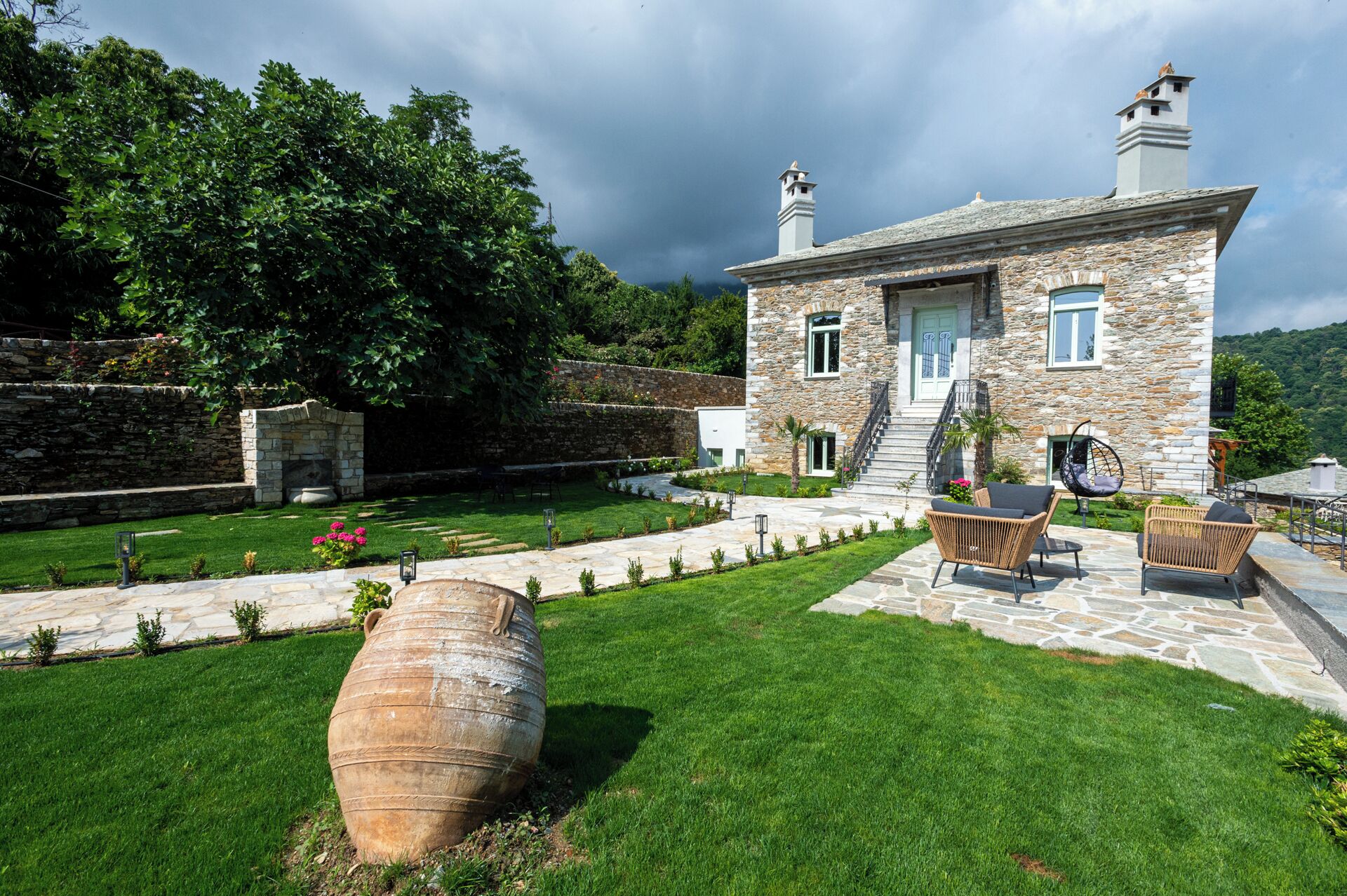 A stone house with a stone staircase leading to a door, surrounded by a well-maintained lawn and a large decorative pot.