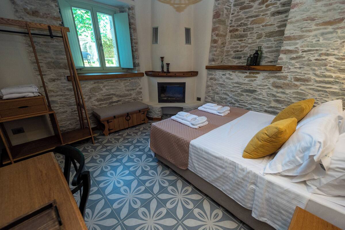 Une chambre avec un mur en pierre, un lit avec des oreillers, un banc, une télévision et une fenêtre avec des volets.