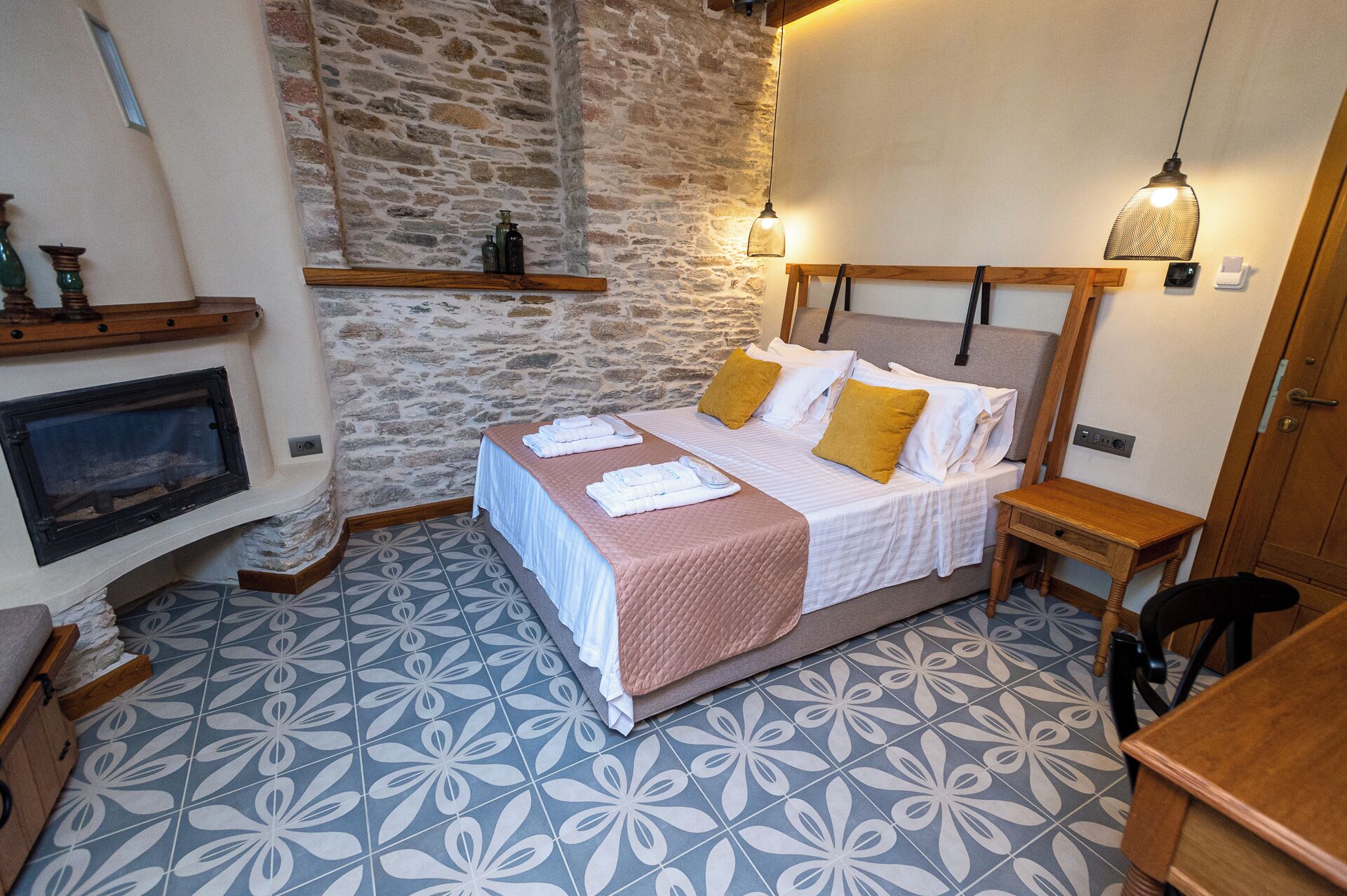 Un dormitorio acogedor con pared de piedra, chimenea, cama con colcha rosa y mesita de noche de madera.