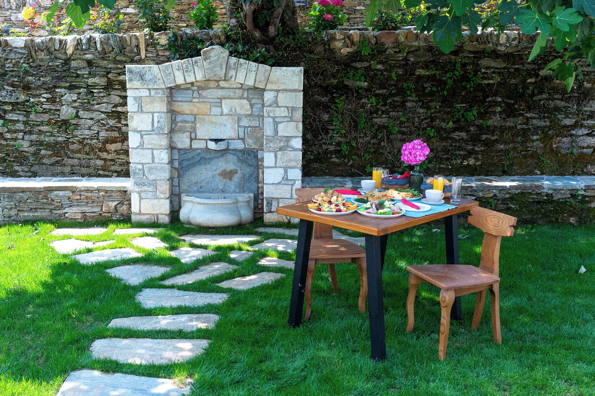 An outdoor dining area with stone steps leading to a table set with plates of food, a stone wall, and a stone fireplace.