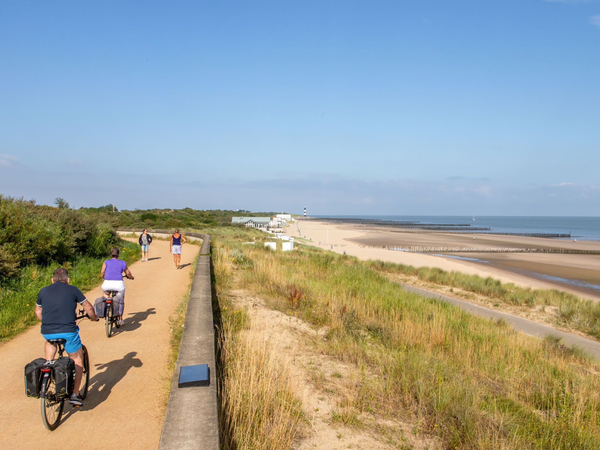 A coastal path with cyclists, a sandy beach, and a lighthouse in the distance.