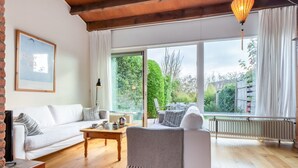Ein modernes Wohnzimmer mit Sofa, Couchtisch und Sessel. Ein großes Fenster bietet Blick auf die grüne Umgebung.