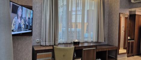 Habitación de hotel con un ventanal grande, un televisor montado en la pared, un escritorio con cajones, una silla y un armario.