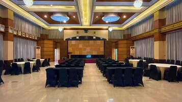 Un amplio salón de conferencias con filas de mesas y sillas, un escenario con un atril y una araña.