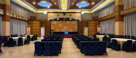 Un amplio salón de conferencias con filas de mesas y sillas, un escenario con un atril y una araña.