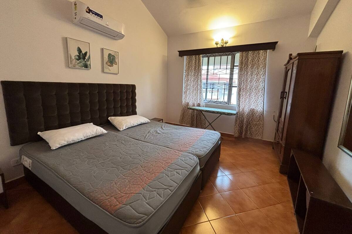 A bedroom with two beds, a ceiling fan, an air conditioning unit, a window with curtains, and a small ironing board.