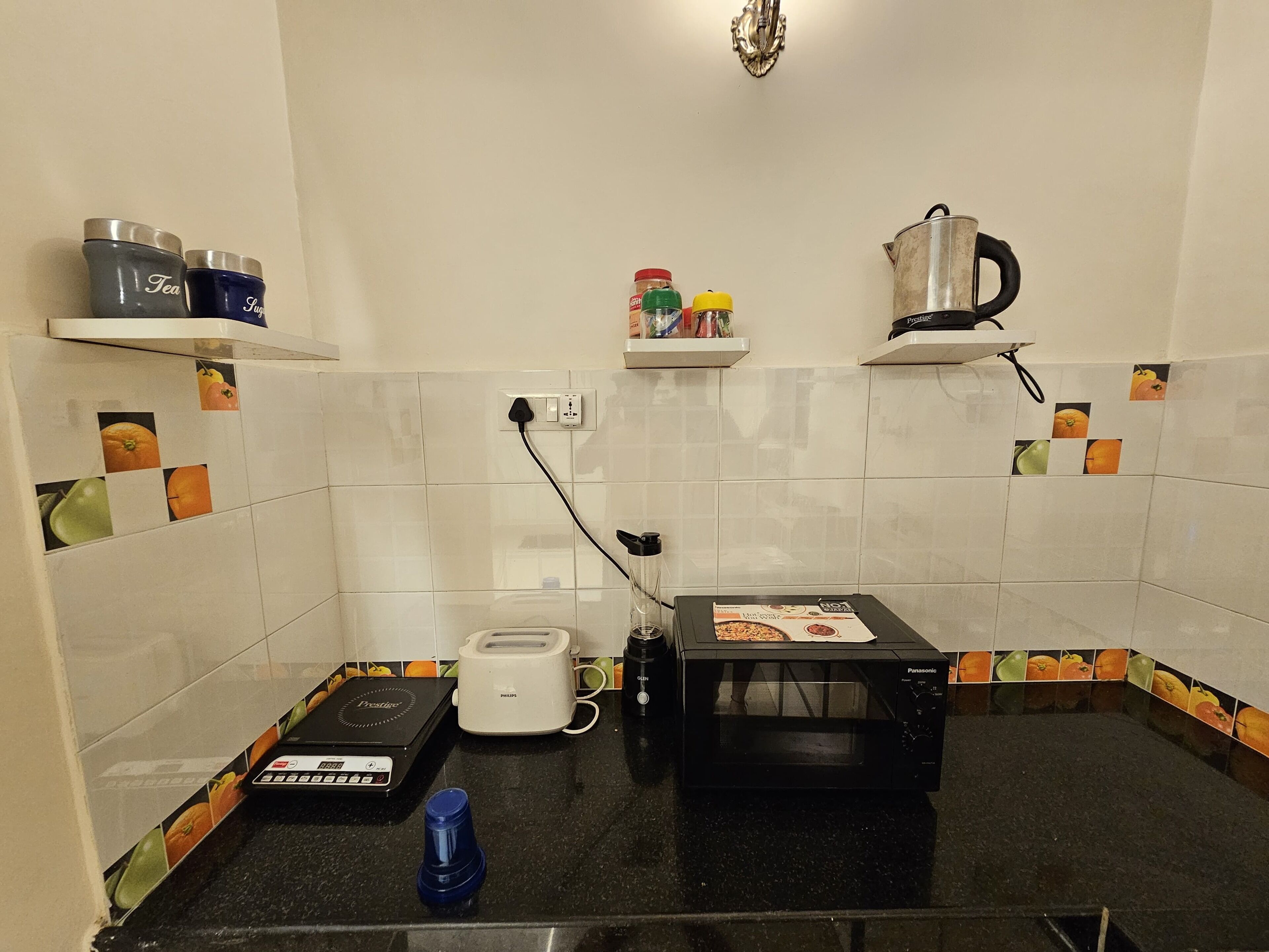 Cocina compacta con microondas, tostadora y hervidor. Dos repisas con frascos y un hervidor en la pared.