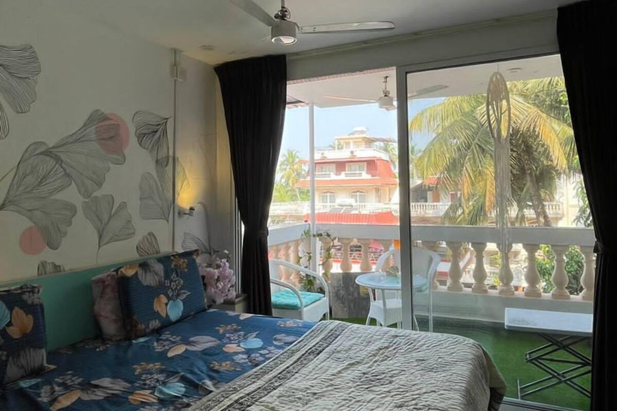 A bedroom with a large bed, a ceiling fan, a balcony with a view of palm trees and buildings.