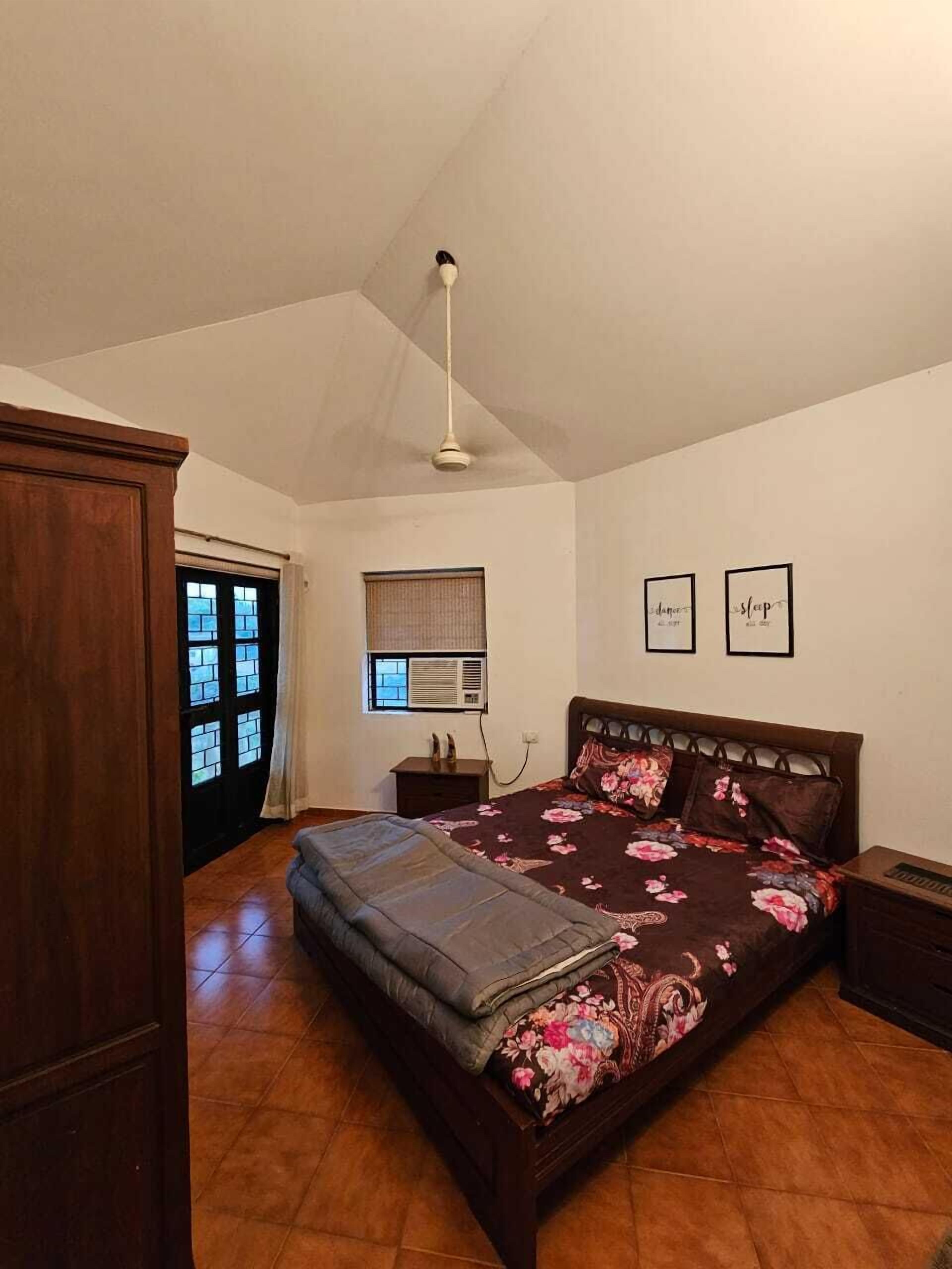 Un dormitorio con una cama grande, un armario de madera, un ventilador de techo, una ventana con persianas y dos cuadros enmarcados en la pared.