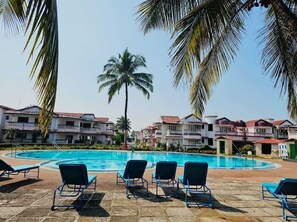 A swimming pool with lounge chairs, palm trees, and multi-story buildings in the background. - Casa De Abhishek - Breezy 2 BHK (Colva)