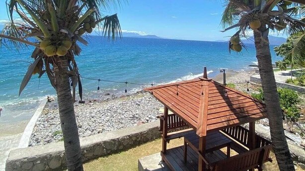 Un tavolo e sedie di legno sotto un tetto verde, con vista su una spiaggia con palme e l'oceano.