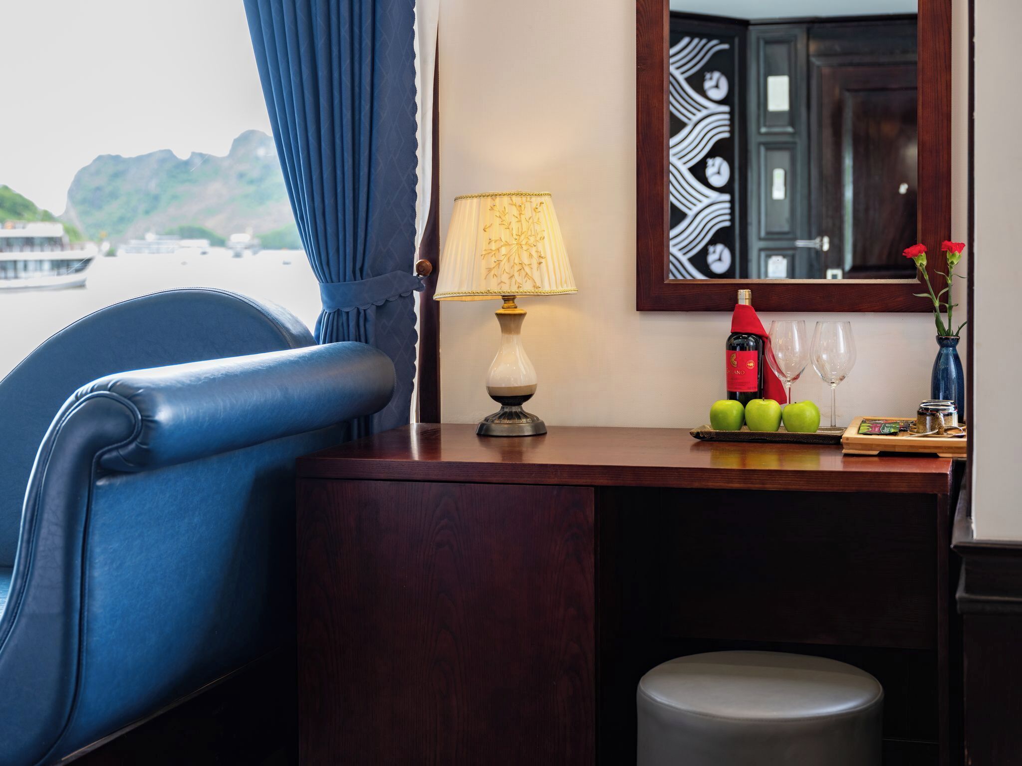 A room with a blue chair, a wooden desk, a lamp, a bottle of wine, two glasses, and a plate with apples.