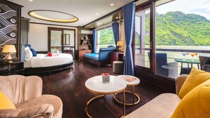 Ein modernes Hotelzimmer mit einem großen Bett, einer Sitzecke mit Sofa und Sesseln, einem kleinen Tisch und einem Balkon mit Blick auf eine üppige grüne Landschaft.