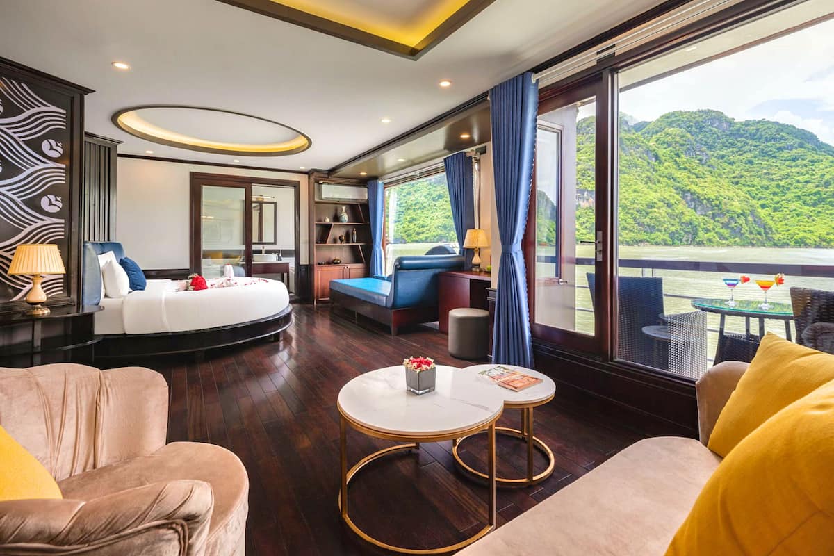 Una habitación de hotel moderna con una cama grande, una zona de estar con un sofá y sillones, una mesita y un balcón con vistas a un paisaje verde y exuberante.