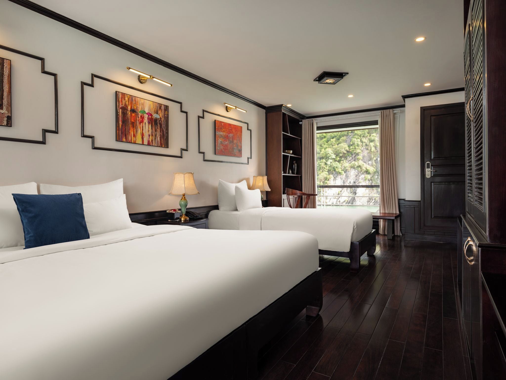 A hotel room with two beds, a bookshelf, and a view of greenery through the window.