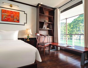 Une chambre d’hôtel avec un grand lit, un bureau, une chaise, une bibliothèque et une fenêtre donnant sur un paysage verdoyant.