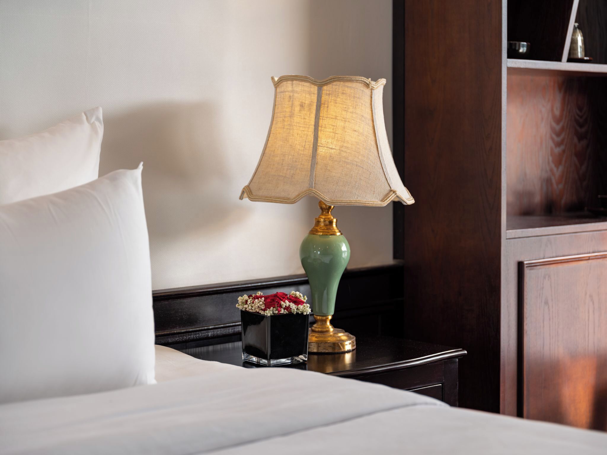 A neatly made bed with white linens, a green lamp with a gold base, and a small vase with red and white flowers on a dark nightstand.