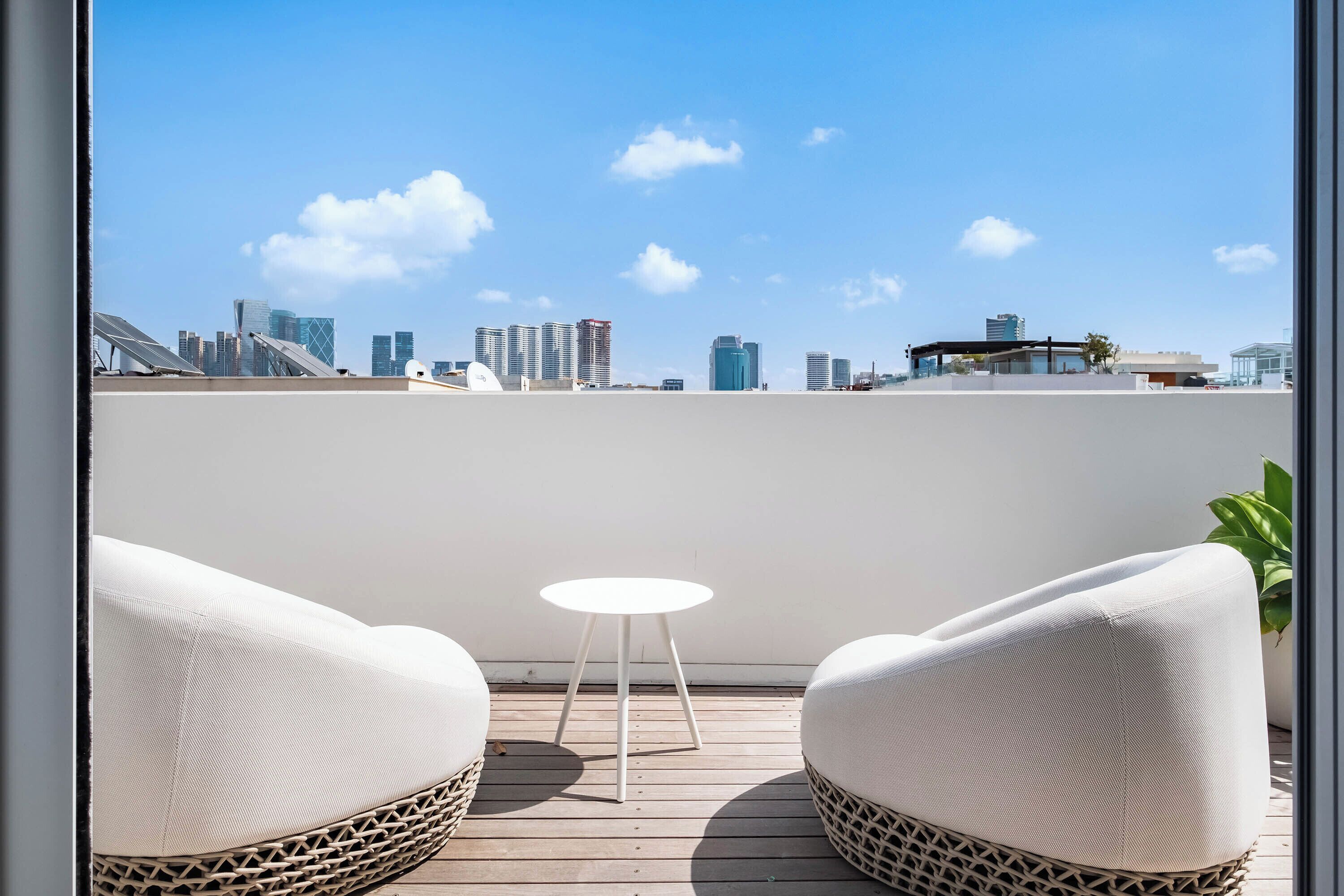 A rooftop terrace with two white cushioned chairs and a small round table, overlooking a city skyline with modern buildings.