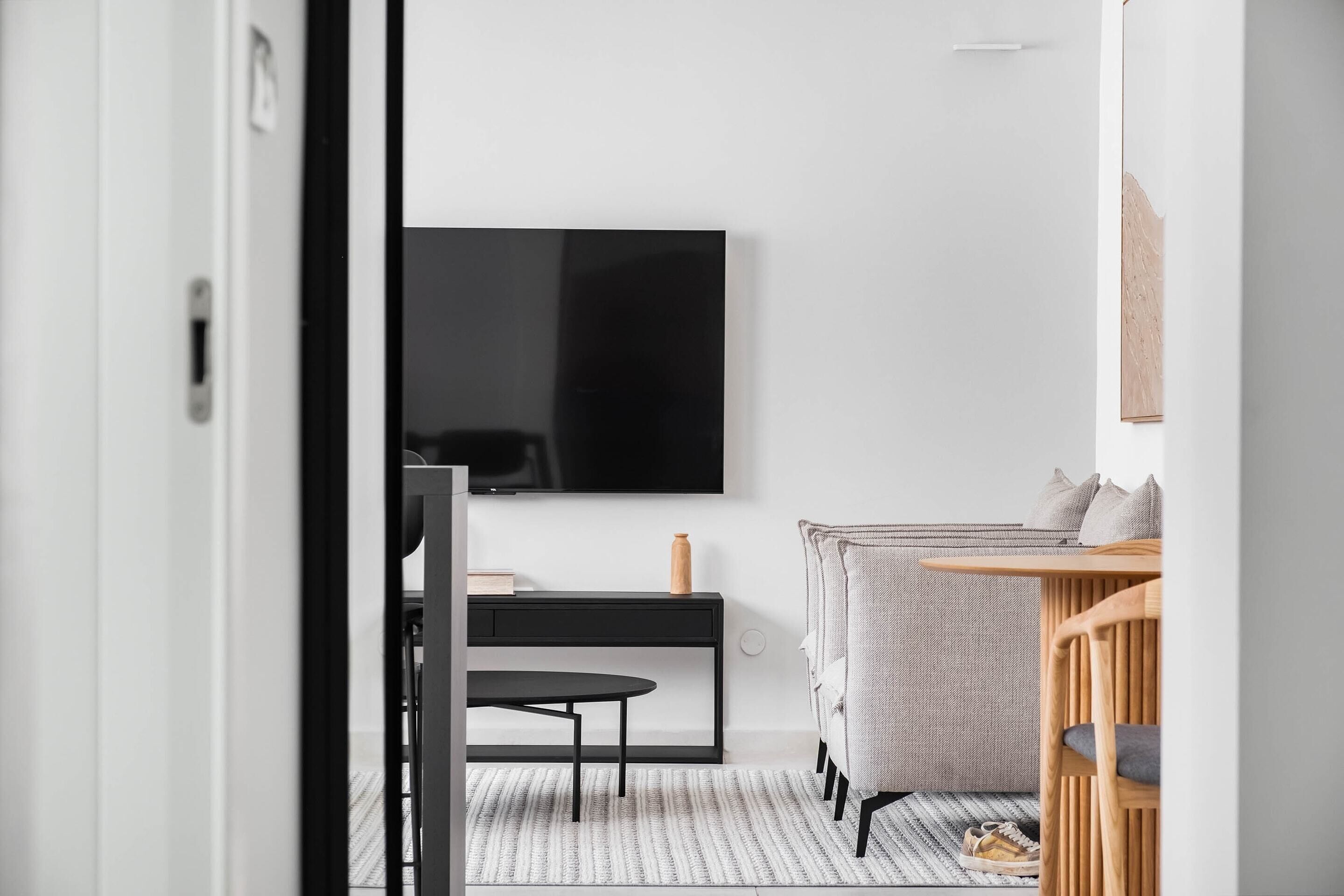 Un salon moderne avec un téléviseur noir fixé au mur, une table d’appoint en bois, un canapé gris et une petite table noire.