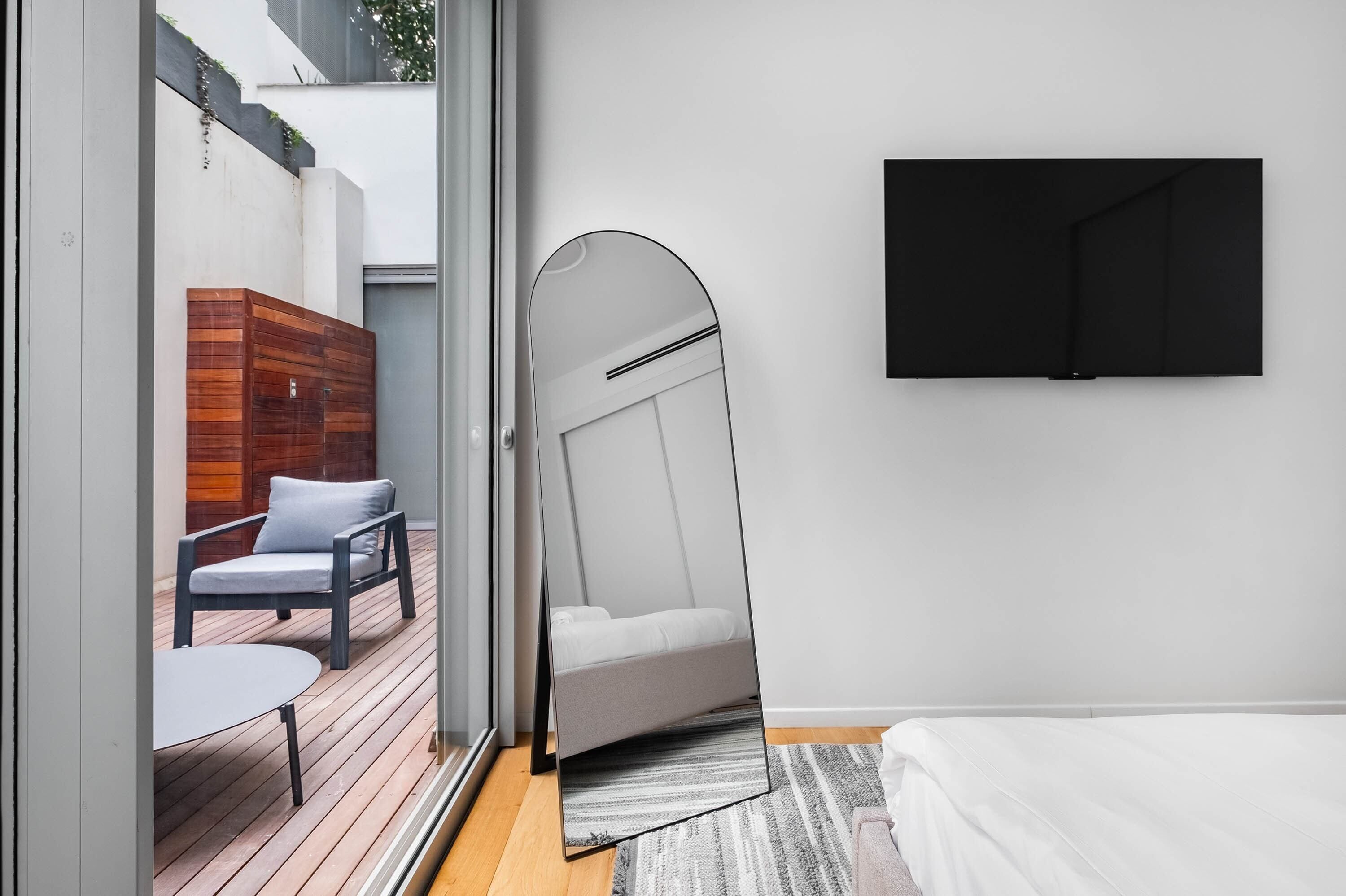 Un dormitorio moderno con una cama grande, un televisor montado en la pared y un ventanal corredero que da a un patio exterior con terraza de madera y zona de estar.