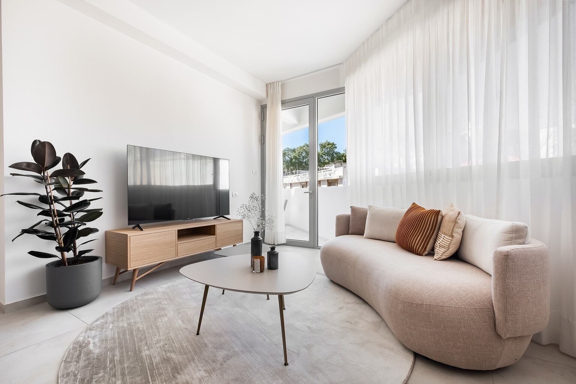 A modern living room with a flat-screen TV, a wooden TV stand, a round coffee table, a beige sofa, and a large window with sheer curtains.