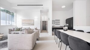 A modern living room with a sofa, a dining area with a long table and chairs, and a kitchen with dark cabinets and a white countertop.