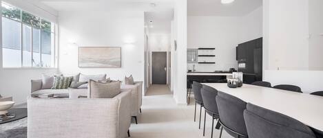 A modern living room with a sofa, a dining area with a long table and chairs, and a kitchen with dark cabinets and a white countertop.