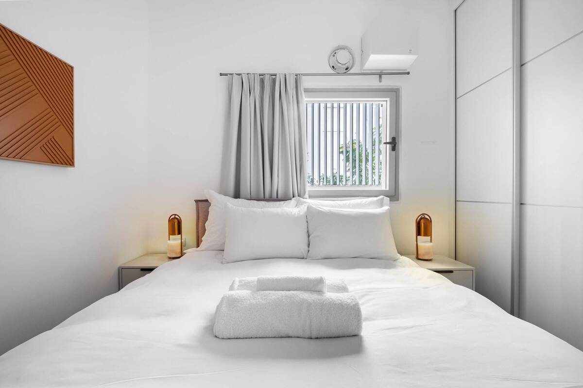 Ein modernes Hotelzimmer mit einem ordentlich bezogenen Bett, zwei Nachttischen mit Lampen, einem Fenster mit Jalousien und einer Wandleuchte.