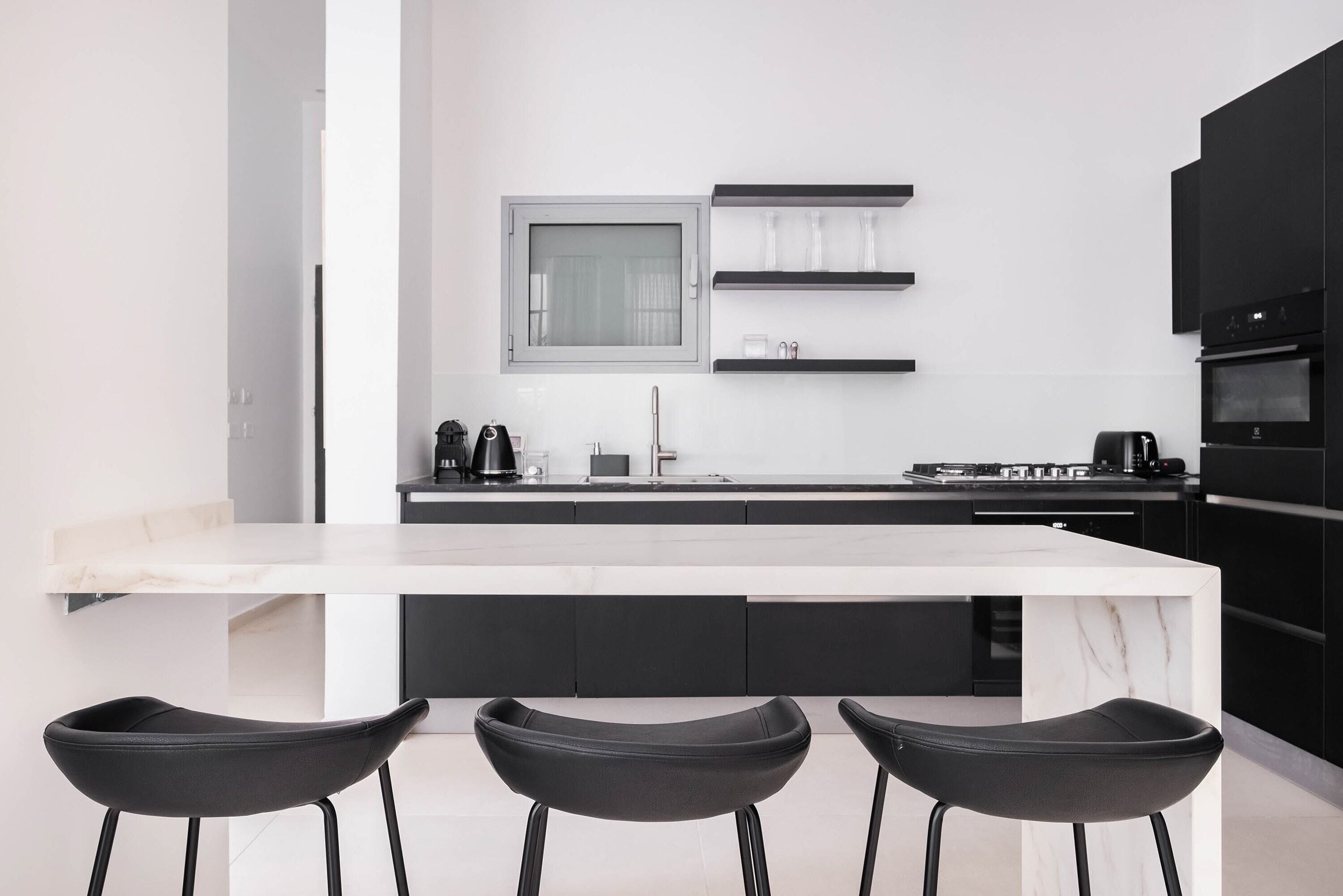 A modern kitchen with a white marble countertop, black cabinets, and three black bar stools.