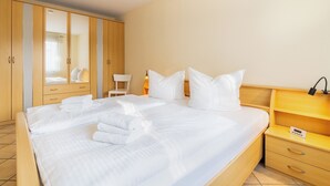 Una cama bien tendida con sábanas y toallas blancas, un armario de madera, una mesita de noche con lámpara y una silla.
