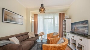Un salon avec un canapé marron, des fauteuils en osier, une petite table ronde surmontée d’un vase, un téléviseur posé sur un meuble et un tableau accroché au mur.